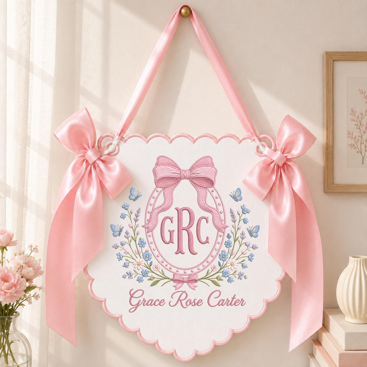 May include: A white scalloped wall hanging featuring pink satin ribbon bows on each side and a top hanging loop. The center displays an embroidered oval frame with a pink bow, the monogram GRC, blue butterflies, and floral accents with the name Grace Rose Carter.