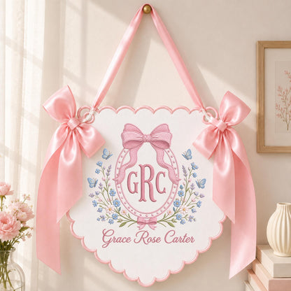May include: A white scalloped wall hanging featuring pink satin ribbon bows on each side and a top hanging loop. The center displays an embroidered oval frame with a pink bow, the monogram GRC, blue butterflies, and floral accents with the name Grace Rose Carter.