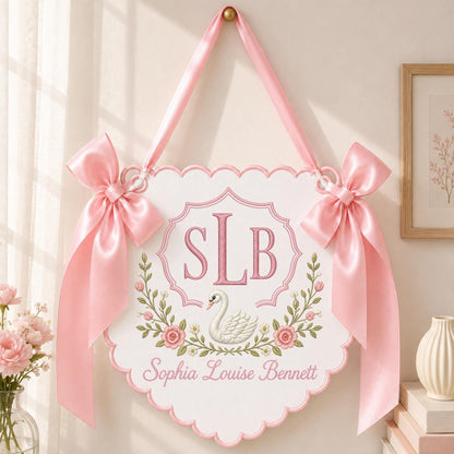 May include: A white scalloped wall hanging featuring a pink embroidered monogram SLB, a white swan illustration, and a floral wreath. The name Sophia Louise Bennett is embroidered in pink below the swan. Two pink satin bows hang from the top.