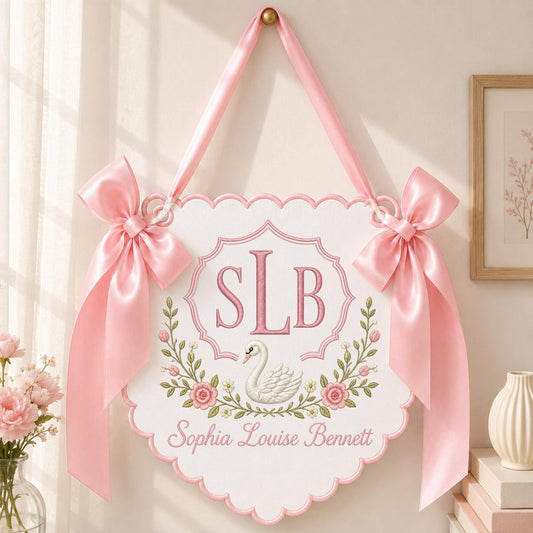 May include: A white scalloped wall hanging featuring a pink embroidered monogram SLB, a white swan illustration, and a floral wreath. The name Sophia Louise Bennett is embroidered in pink below the swan. Two pink satin bows hang from the top.
