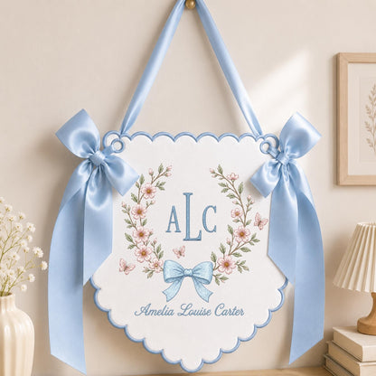 May include: A white scalloped wall hanging featuring a blue embroidered monogram ALC surrounded by a floral wreath with pink flowers and small butterflies. A blue satin bow is at the bottom, and two blue satin ribbons are tied at the top corners.