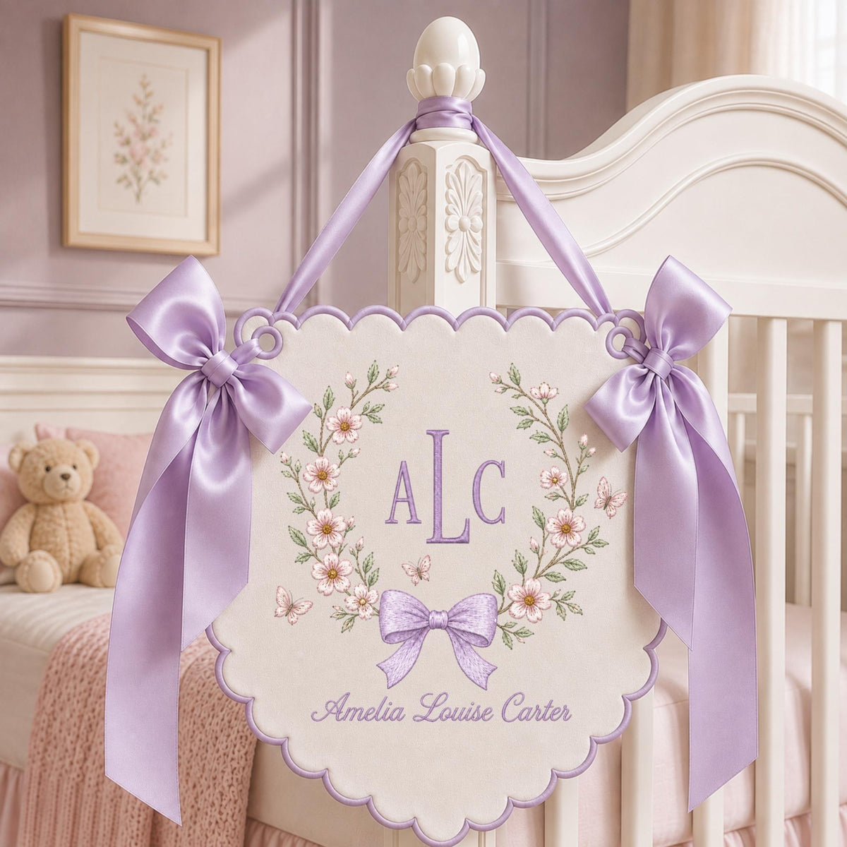 May include: A cream-colored scalloped wall hanging featuring a floral wreath, butterflies, and a purple bow. The center displays the monogram ALC, with the name Amelia Louise Carter printed below. Two large lavender satin ribbons hang from the top.