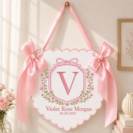 May include: A white scalloped wall hanging featuring a pink embroidered letter V inside a floral wreath. The sign is personalized with the name Violet Rose Morgan and the date 10.20.2027, accented by two large pink satin bows and a matching ribbon hanger.