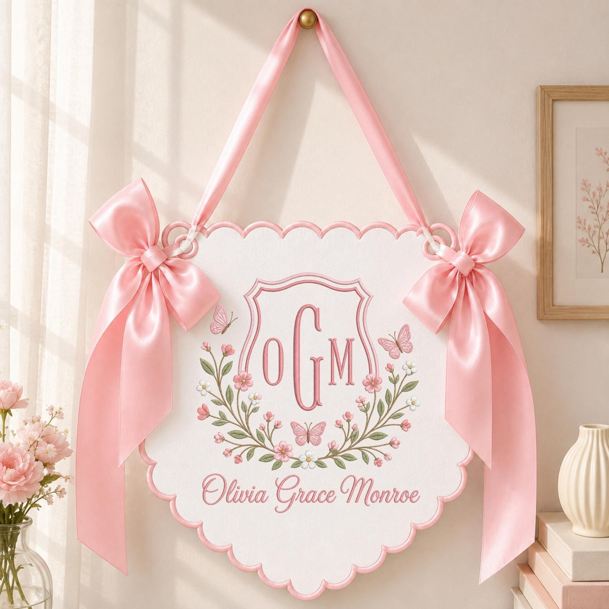 May include: A white scalloped wall hanging featuring a pink embroidered shield with the monogram OGM, surrounded by floral vines and butterflies. The sign includes the name Olivia Grace Monroe in pink script and two large pink satin bows.