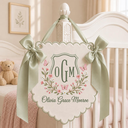 May include: A cream-colored scalloped wall hanging featuring sage green embroidery of the monogram OGM, pink flowers, and butterflies. The sign includes the name Olivia Grace Monroe in sage green script and is adorned with two sage green satin bows.