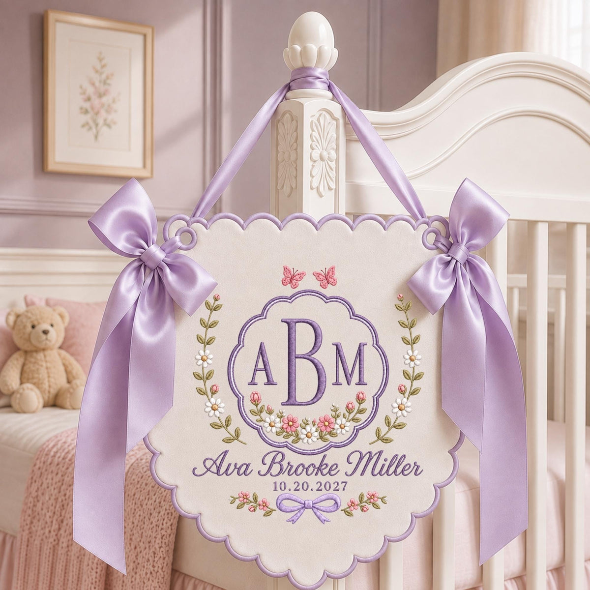 May include: A cream-colored fabric wall hanging with scalloped edges, featuring purple embroidered monogram letters ABM, the name Ava Brooke Miller, and the date 10.20.2027. It is decorated with floral vines, butterflies, and two large lavender satin bows.