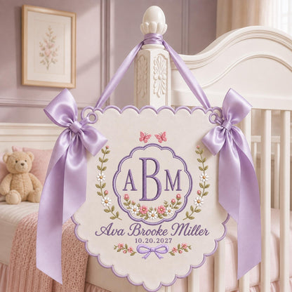 May include: A cream-colored fabric wall hanging with scalloped edges, featuring purple embroidered monogram letters ABM, the name Ava Brooke Miller, and the date 10.20.2027. It is decorated with floral vines, butterflies, and two large lavender satin bows.