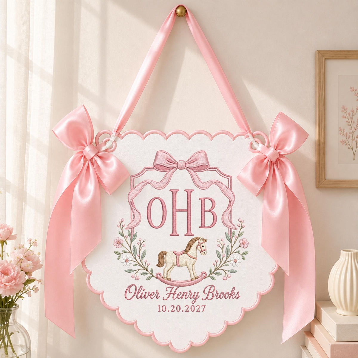 May include: A white scalloped wall hanging featuring pink satin bows on each side and a pink ribbon for hanging. The center displays the embroidered monogram OHB, a rocking horse illustration, and the text Oliver Henry Brooks 10.20.2027.