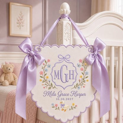 May include: A cream-colored scalloped wall hanging featuring purple embroidered monogram letters MGH, the name Mila Grace Harper, and the date 10.20.2027. The design includes floral motifs, butterflies, and a duck, accented by two large lavender satin bows.