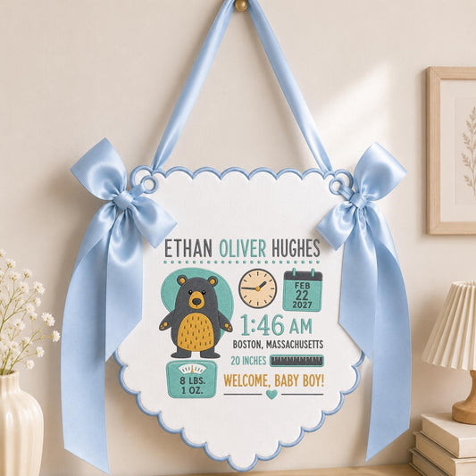 May include: A white scalloped wall hanging with light blue ribbon bows, featuring a bear illustration and text: ETHAN OLIVER HUGHES, FEB 22 2027, 1:46 AM, BOSTON, MASSACHUSETTS, 20 INCHES, 8 LBS. 1 OZ., and WELCOME, BABY BOY! with a clock and calendar.