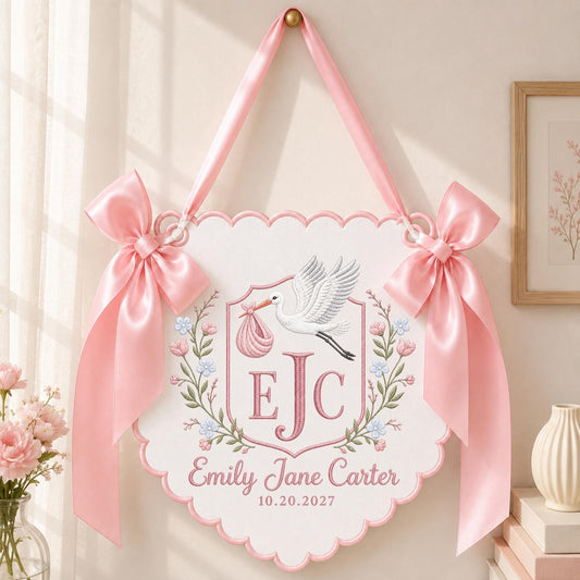 May include: A white scalloped wall hanging featuring a pink embroidered stork carrying a bundle, the initials EJC, and the text Emily Jane Carter 10.20.2027. The piece is decorated with floral embroidery and two large pink satin bows on each side.