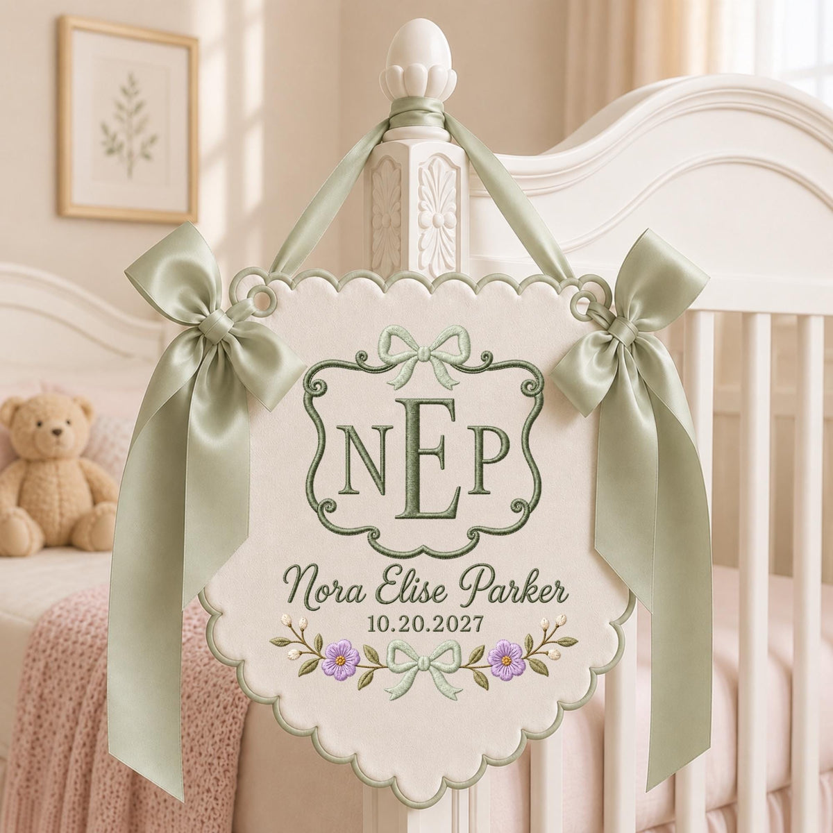 May include: A cream-colored fabric wall hanging with scalloped edges, featuring sage green embroidery of the monogram NEP, the name Nora Elise Parker, and the date 10.20.2027. It is adorned with two sage green satin bows and a matching ribbon hanger.
