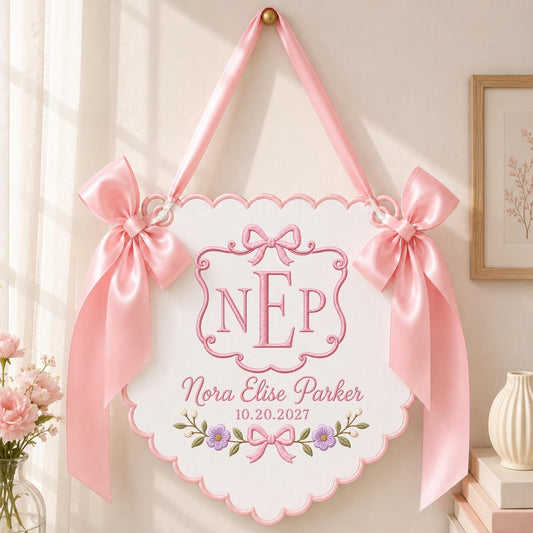 May include: A white scalloped wall hanging featuring pink embroidered details, including the monogram NEP, the name Nora Elise Parker, the date 10.20.2027, and floral accents. The piece is suspended by a pink ribbon and adorned with two large pink bows.