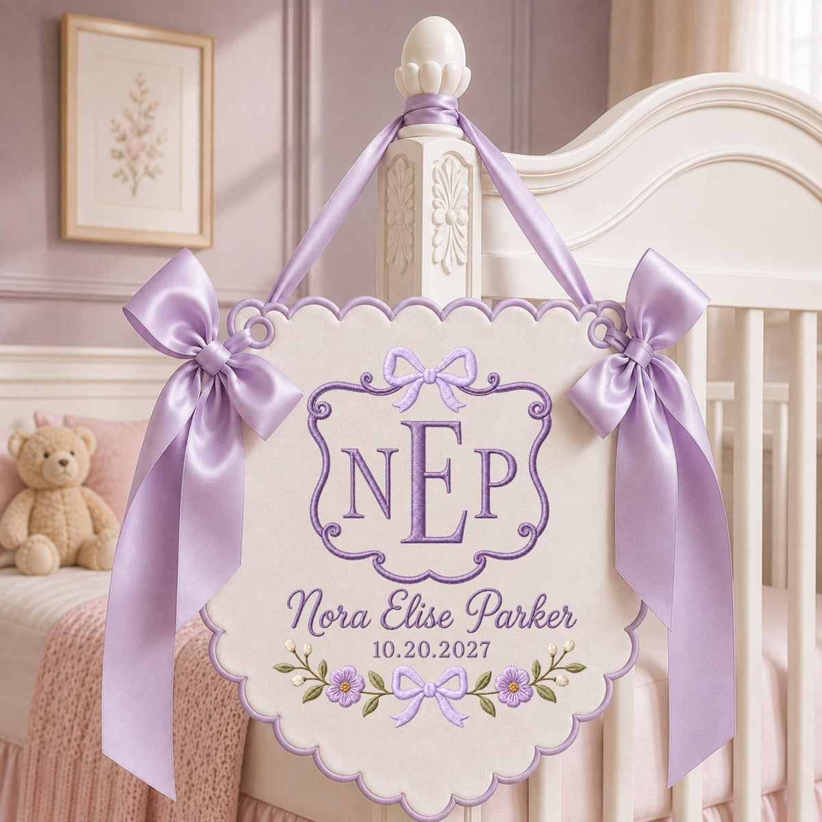 May include: A cream-colored scalloped wall hanging featuring purple embroidery of the monogram NEP, the name Nora Elise Parker, and the date 10.20.2027. The banner is decorated with purple floral motifs and two large lavender satin ribbon bows.