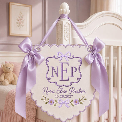 May include: A cream-colored scalloped wall hanging featuring purple embroidery of the monogram NEP, the name Nora Elise Parker, and the date 10.20.2027. The banner is decorated with purple floral motifs and two large lavender satin ribbon bows.