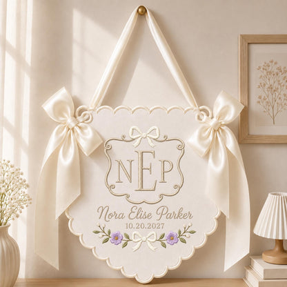 May include: A cream-colored scalloped wall hanging featuring an embroidered monogram NEP, the name Nora Elise Parker, and the date 10.20.2027. The piece is adorned with two large satin bows on the sides and a matching ribbon for hanging.