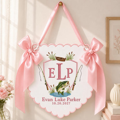 May include: A white scalloped wall hanging with pink satin bows on each side. The center features a pink shield with the monogram ELP, a small boat, two fishing rods, and a green fish. Below, the text reads Evan Luke Parker and 10.20.2027.