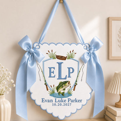 May include: A white scalloped wall hanging featuring a light blue embroidered shield with the letters ELP, a brown boat, two fishing rods, and a green fish. The text reads Evan Luke Parker and 10.20.2027, accented by two light blue satin bows.