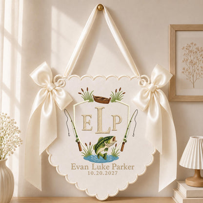 May include: A cream-colored scalloped wall hanging featuring an embroidered design with the monogram ELP, a brown boat, two fishing rods, and a green fish. The text reads Evan Luke Parker and 10.20.2027. It is accented with two large satin bows.