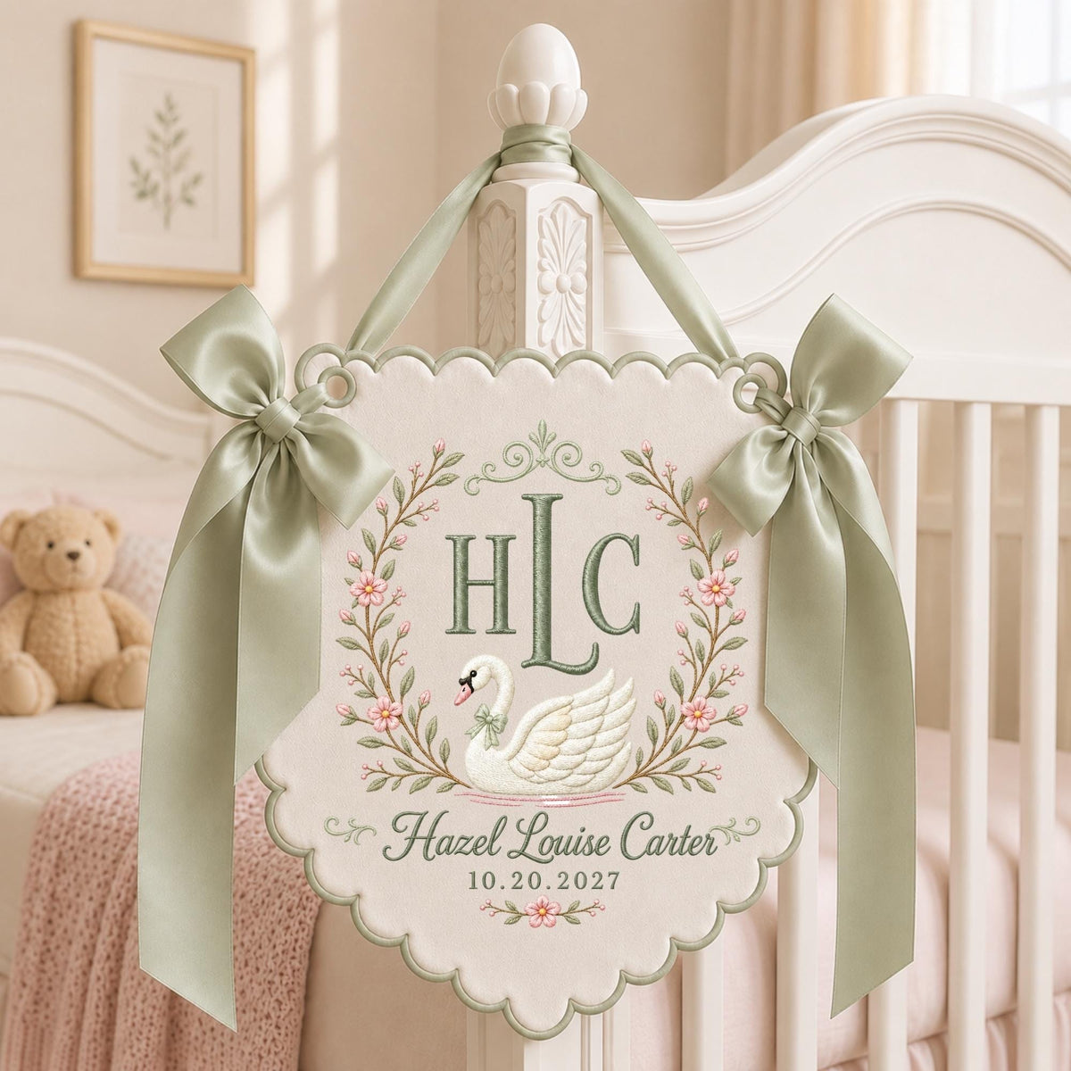 May include: A cream-colored scalloped wall hanging featuring an embroidered white swan, a floral wreath, and the monogram HLC. The text reads Hazel Louise Carter and 10.20.2027. It is decorated with two sage green satin ribbons tied in bows.