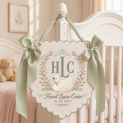 May include: A cream-colored scalloped wall hanging featuring an embroidered white swan, a floral wreath, and the monogram HLC. The text reads Hazel Louise Carter and 10.20.2027. It is decorated with two sage green satin ribbons tied in bows.
