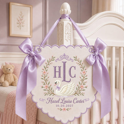 May include: A scalloped, off-white fabric wall hanging featuring an embroidered swan, floral wreath, and the monogram HLC. The text reads Hazel Louise Carter and 10.20.2027. It is adorned with two large, light purple satin bows and matching ribbon.