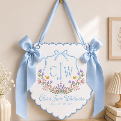 May include: A white scalloped wall hanging featuring light blue satin ribbon bows on each side. The embroidery includes the monogram CJW, a floral wreath with lavender and small yellow butterflies, and the text Clara June Whitmore 07.16.2027.