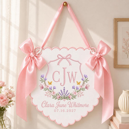 May include: A white scalloped wall hanging featuring a pink embroidered monogram CJW, floral wreath, and the text Clara June Whitmore 07.16.2027. The sign is suspended by a pink satin ribbon and decorated with two large pink satin bows on each side.