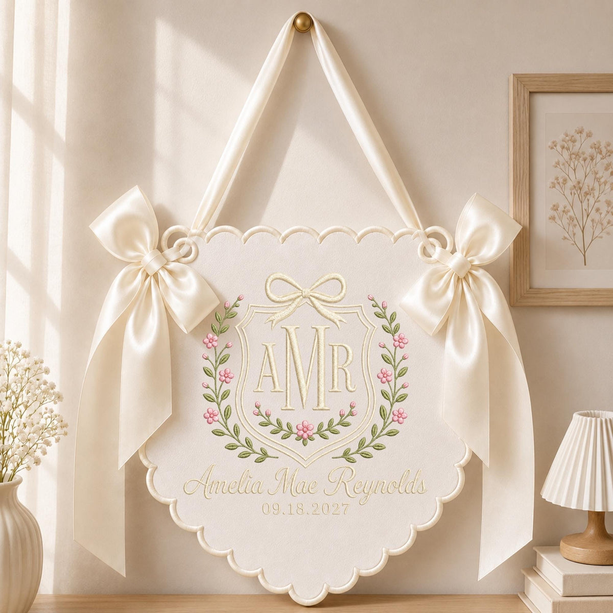 May include: A cream-colored scalloped wall hanging featuring a central embroidered crest with the initials AMR, a bow, and floral vines. The text Amelia Mae Reynolds and 09.18.2027 is embroidered below. Two large satin ribbons are tied at the top sides.