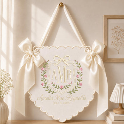 May include: A cream-colored scalloped wall hanging featuring a central embroidered crest with the initials AMR, a bow, and floral vines. The text Amelia Mae Reynolds and 09.18.2027 is embroidered below. Two large satin ribbons are tied at the top sides.