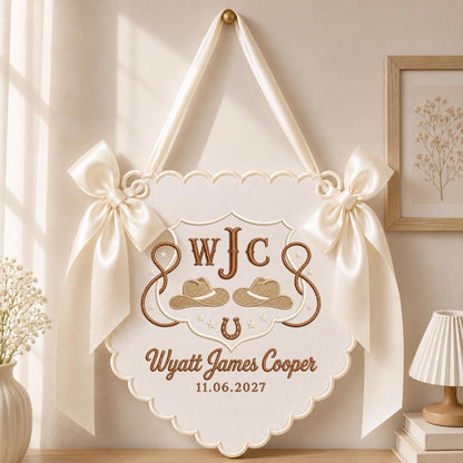 May include: A cream-colored scalloped wall hanging featuring brown embroidery of the monogram WJC, two cowboy hats, a horseshoe, and a lasso. The text reads Wyatt James Cooper and 11.06.2027. It is adorned with two large satin ribbons on the sides.