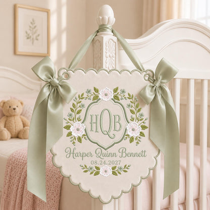 May include: A cream-colored fabric wall hanging with scalloped edges, featuring sage green and pink floral embroidery. The center displays the monogram HQB, the name Harper Quinn Bennett, and the date 08.24.2027, accented by two sage green satin bows.