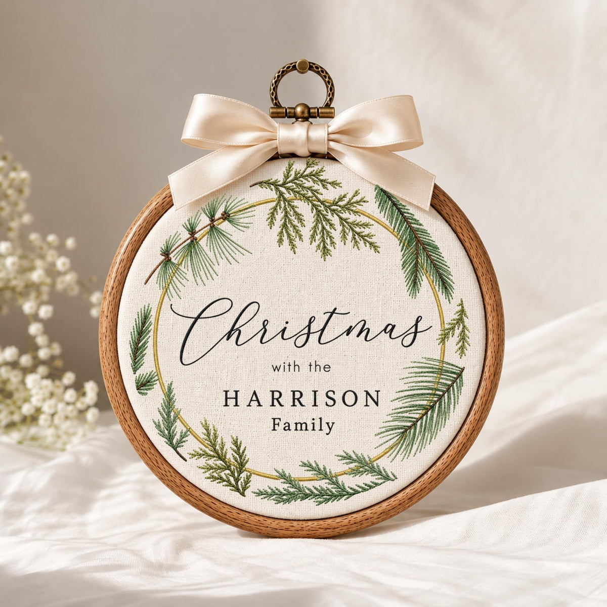 May include: A round wooden embroidery hoop containing off-white fabric with a printed green pine wreath design. The text reads "Christmas with the HARRISON Family" in black script and serif fonts. A cream-colored satin ribbon bow is attached at the top.