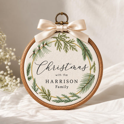 May include: A round wooden embroidery hoop containing off-white fabric with a printed green pine wreath design. The text reads "Christmas with the HARRISON Family" in black script and serif fonts. A cream-colored satin ribbon bow is attached at the top.