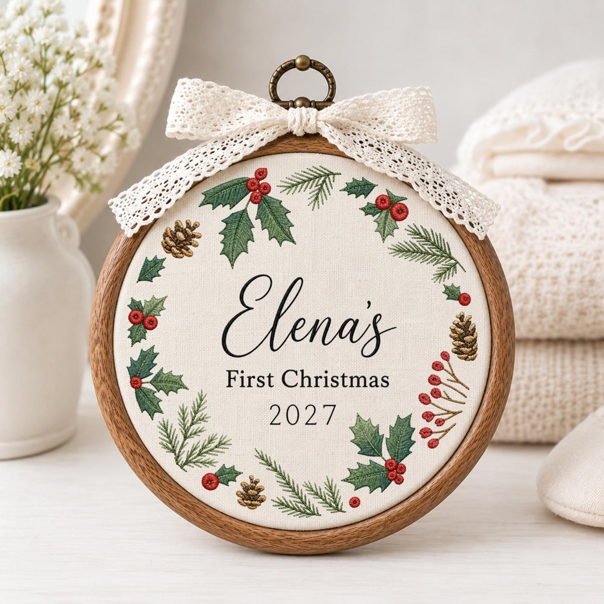May include: A circular wooden embroidery hoop frame containing off-white fabric with a printed wreath of green holly leaves, red berries, and brown pinecones. The text reads Elena's First Christmas 2027. A cream lace bow is attached to the top.