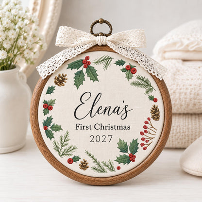 May include: A circular wooden embroidery hoop frame containing off-white fabric with a printed wreath of green holly leaves, red berries, and brown pinecones. The text reads Elena's First Christmas 2027. A cream lace bow is attached to the top.