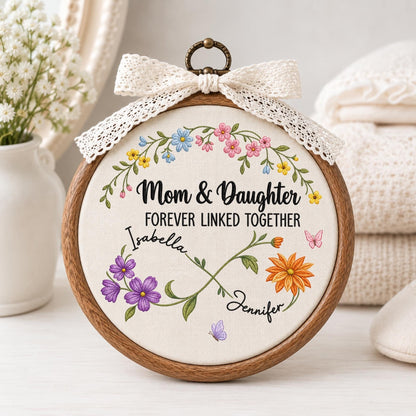May include: A circular wooden embroidery hoop containing white fabric with floral embroidery. The text reads Mom &amp; Daughter FOREVER LINKED TOGETHER with the names Isabella and Jennifer. An infinity symbol is formed by stems with purple and orange flowers.