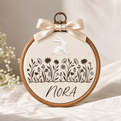 May include: A circular wooden embroidery hoop containing cream-colored fabric with a white stitched rabbit, dark brown embroidered wildflowers, and the name NORA in dark brown script. A beige satin ribbon bow is attached to the top metal hanger.