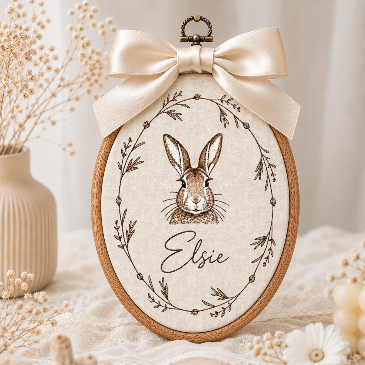 May include: An oval embroidery hoop featuring a cream-colored fabric panel with a brown line-art illustration of a rabbit centered inside a delicate botanical wreath. The name Elsie is embroidered below the rabbit. A large cream satin bow is attached.