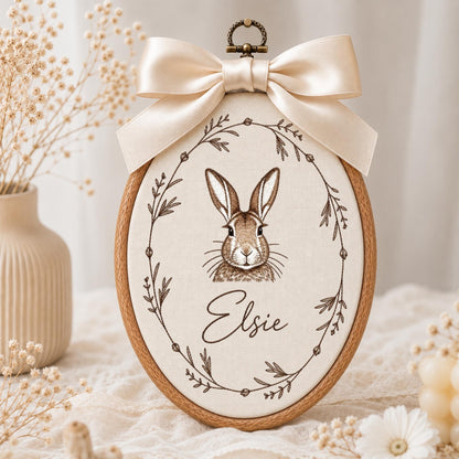 May include: An oval embroidery hoop featuring a cream-colored fabric panel with a brown line-art illustration of a rabbit centered inside a delicate botanical wreath. The name Elsie is embroidered below the rabbit. A large cream satin bow is attached.