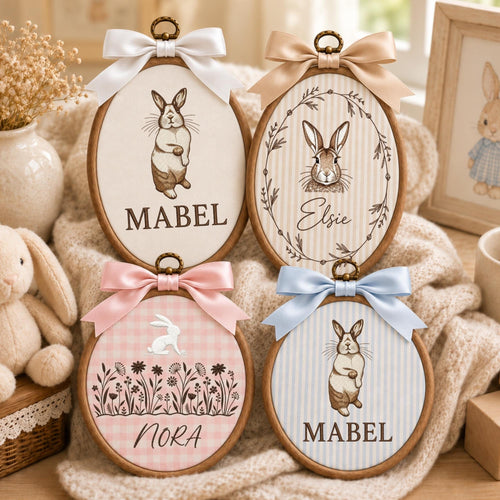 May include: Four oval wooden embroidery hoops featuring rabbit illustrations and names. The top left shows a rabbit and the name MABEL with a white bow. The top right shows a rabbit in a wreath and the name Elsie with a tan bow. The bottom left shows a rabbit, flowers, and the name NORA with a pink bow. The bottom right shows a rabbit and the name MABEL with a light blue bow.