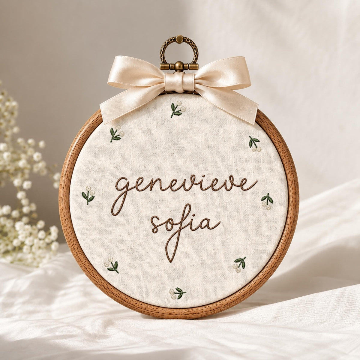May include: A circular wooden embroidery hoop containing off-white fabric with the embroidered brown text "genevieve sofia" in a cursive script, surrounded by small green and white floral sprigs, topped with a cream-colored satin ribbon bow.