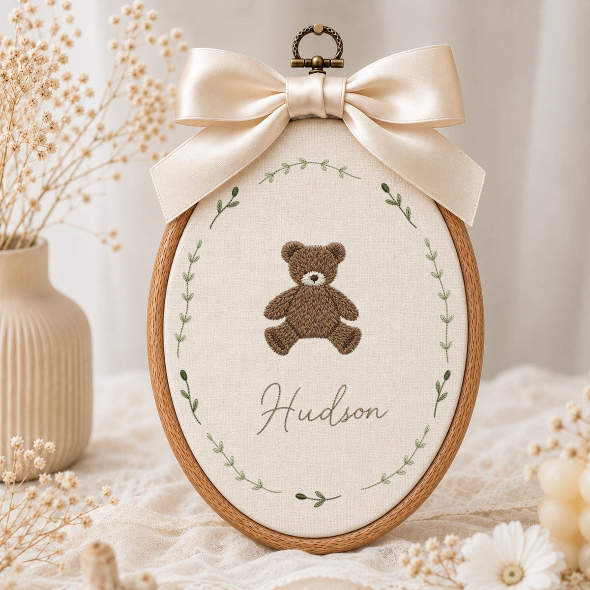 May include: An oval wooden embroidery hoop containing cream-colored fabric with a brown teddy bear and the name Hudson embroidered in the center. A green leaf wreath surrounds the bear, and a large cream satin ribbon is tied at the top.