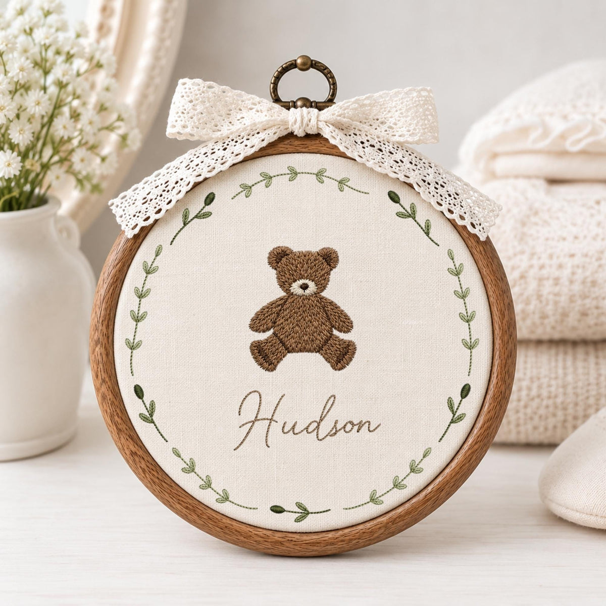 May include: A circular wooden embroidery hoop containing off-white fabric with a brown teddy bear stitched in the center. The name Hudson is embroidered below the bear in cursive script, surrounded by a green leaf wreath and a white lace bow.