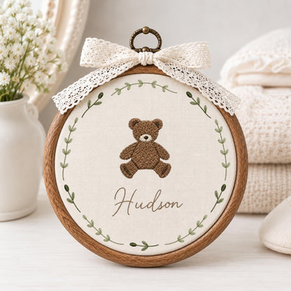 May include: A circular wooden embroidery hoop containing off-white fabric with a brown teddy bear stitched in the center. The name Hudson is embroidered below the bear in cursive script, surrounded by a green leaf wreath and a white lace bow.