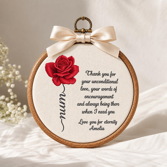 May include: A circular wooden embroidery hoop containing off-white fabric with a red rose illustration, the word mum in cursive, and black text reading Thank you for your unconditional love, your words of encouragement and always being there when I need you. Love you for eternity Amelia. A cream satin ribbon bow is attached to the top.