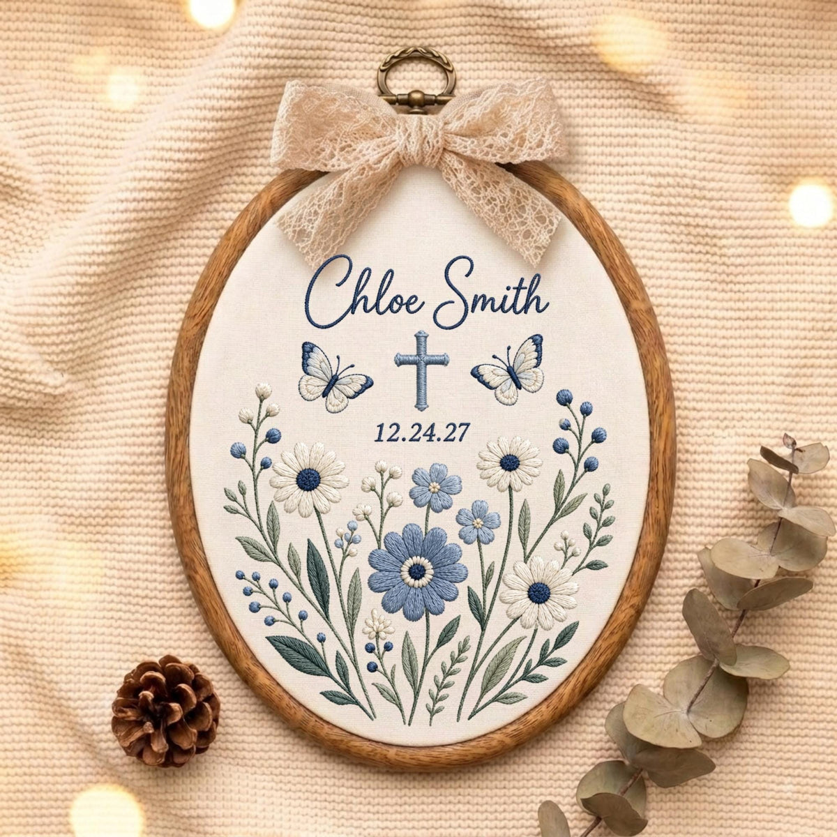 May include: An oval wooden embroidery hoop containing white fabric with blue floral embroidery, two butterflies, a cross, and the text Chloe Smith 12.24.27. A beige lace bow is attached to the top of the hoop, which features a metal hanging loop.