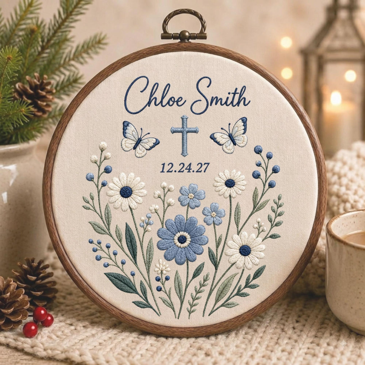 May include: A circular wooden embroidery hoop containing cream fabric with blue thread embroidery. The design features the text Chloe Smith, a cross, two butterflies, and a floral arrangement of blue and white daisies and green leaves with the date 12.24.27.