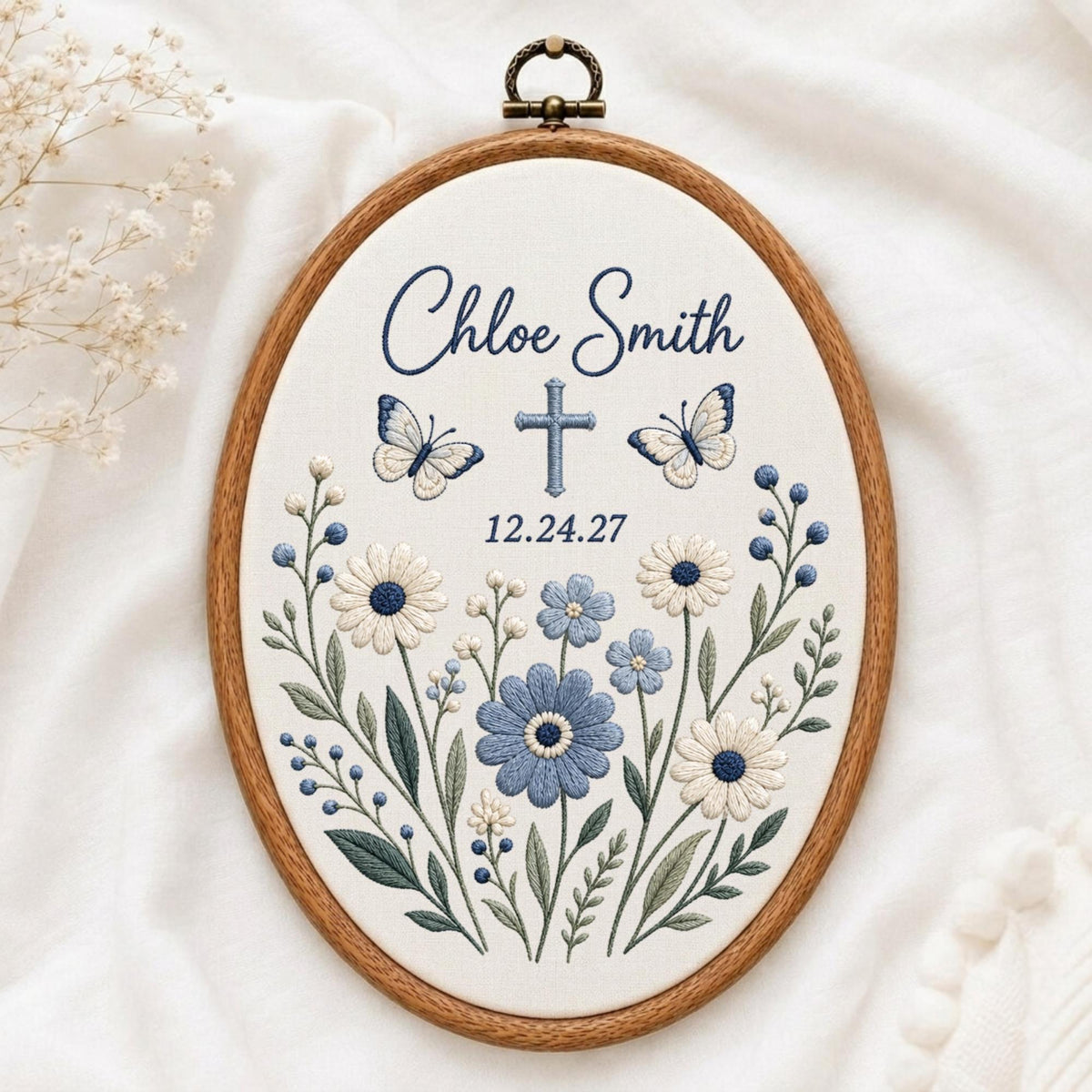 May include: An oval wooden embroidery hoop containing white fabric with blue and white floral embroidery. The design features the text Chloe Smith, a cross, the date 12.24.27, and two butterflies. The embroidery uses shades of blue and white thread.