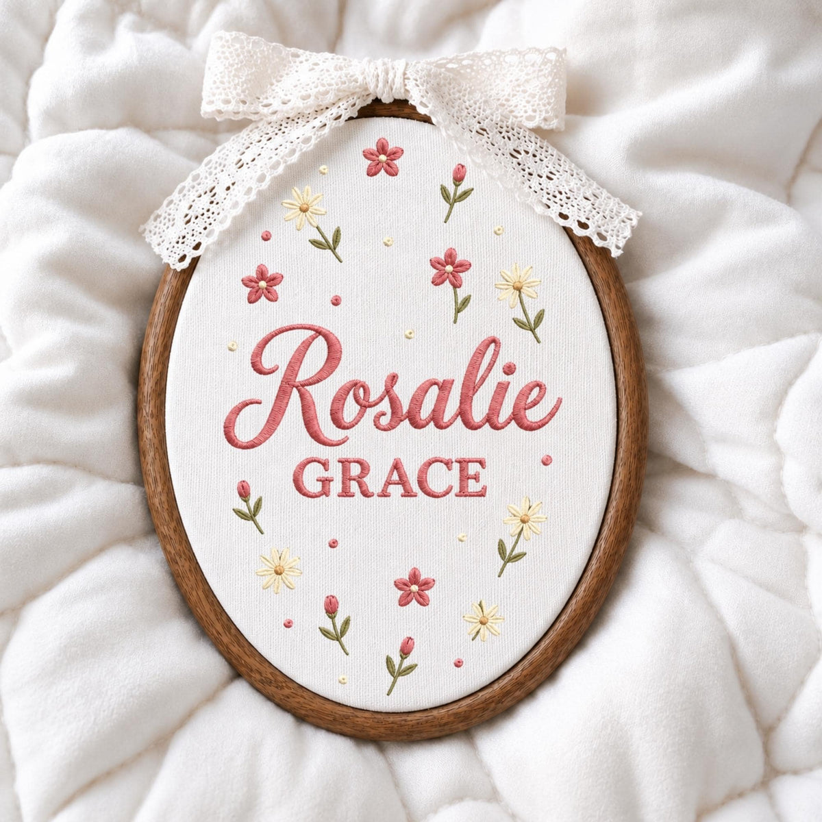 May include: An oval wooden embroidery hoop containing white fabric with the embroidered name Rosalie GRACE in pink script and block lettering. The design features small pink and yellow flowers with green stems and a white lace bow at the top.
